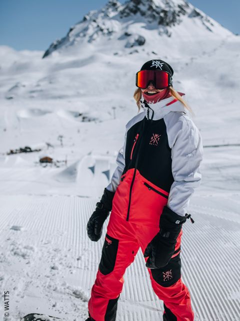 Una persona con un traje de esquí rojo y negro está de pie sonriendo en una montaña nevada y lleva gafas de esquí, guantes y un gorro en un día soleado de invierno.