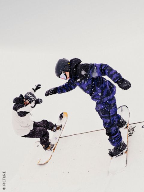 Dos snowboarders con equipo de invierno realizan trucos en una rampa nevada, rodeados de nieve que cae bajo un cielo cubierto.