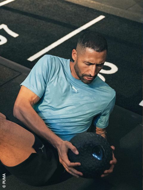 Un hombre con una camiseta Puma azul claro se ejercita en el suelo de un gimnasio y sostiene con ambas manos un balón medicinal negro texturizado.