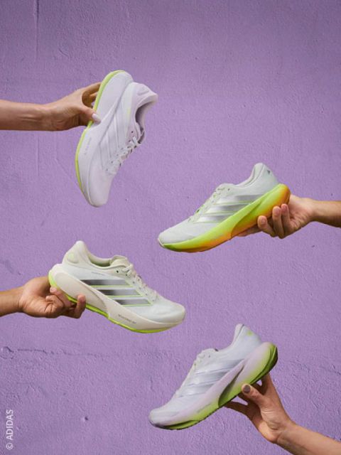 Cuatro manos que sostienen zapatillas blancas con suelas verdes y amarillas frente a una pared de color lavanda.