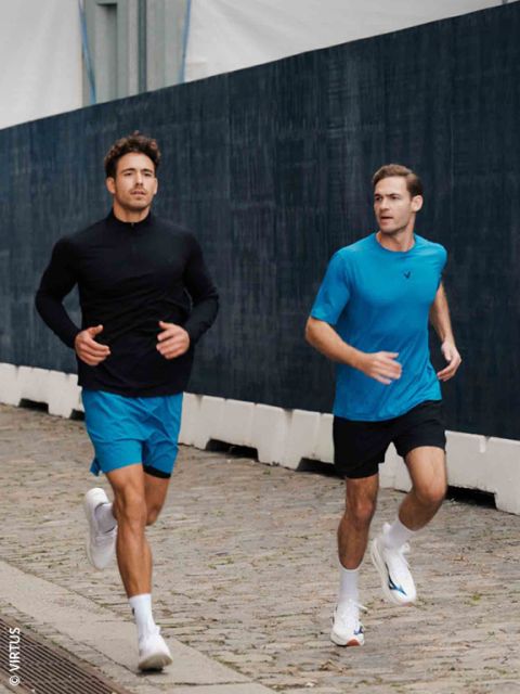 Dos hombres con ropa deportiva trotan por un camino empedrado junto a una valla oscura; ambos llevan shorts azules y zapatillas blancas.