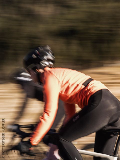 Un ciclista, que lleva una chaqueta verde Löffler y un casco, mira hacia adelante con una mirada concentrada. La imagen está ligeramente borrosa, lo que enfatiza los detalles de la parte superior del cuerpo y la chaqueta.