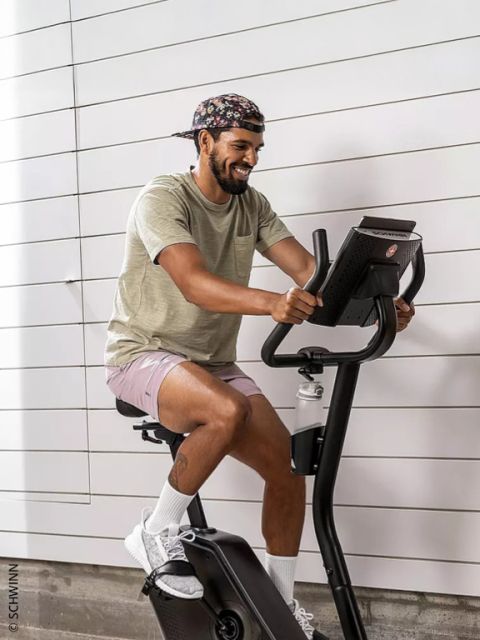Un hombre con una gorra floreada, una camiseta beige y pantalones cortos rosas sonríe mientras monta en una bicicleta estática Schwinn.