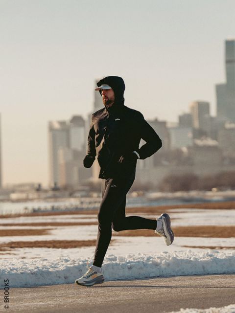 Una persona con una sudadera con capucha y zapatillas de correr trota por un sendero nevado en un día frío y despejado, con el horizonte de la ciudad al fondo.