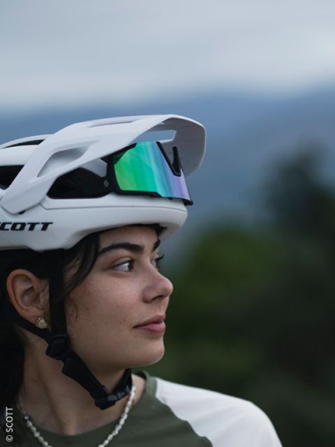 Una joven con un casco de bicicleta blanco y gafas de sol reflectantes en la cabeza mira hacia un lado. El fondo está borroso con vegetación natural y un cielo suave y nublado.
