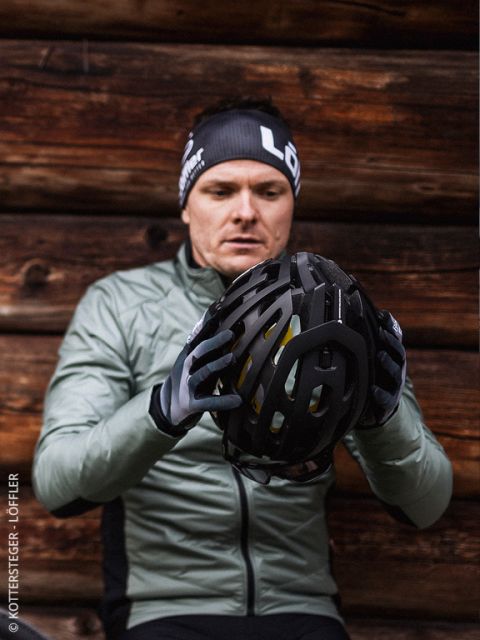 Un hombre con una chaqueta verde y guantes examina un casco de bicicleta negro y está sentado frente a una pared de madera. Lleva una cinta negra para la cabeza con letras blancas.