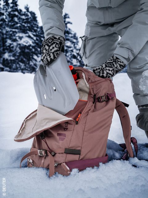 Una persona con ropa de invierno y guantes estampados pone una pala de nieve en una mochila rosa sobre un suelo cubierto de nieve, con árboles nevados al fondo.