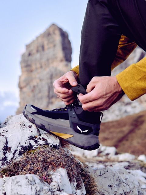 Una persona con ropa de exterior se ata los cordones de una bota de montaña negra mientras está de pie sobre una superficie rocosa, con una gran formación rocosa y un cielo azul de fondo.