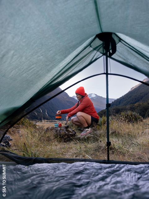 Una persona con chaqueta y gorro rojos prepara comida al aire libre, sentada en la hierba, cerca de equipo de camping, vista a través de la puerta abierta de una tienda de campaña con montañas y árboles al fondo.