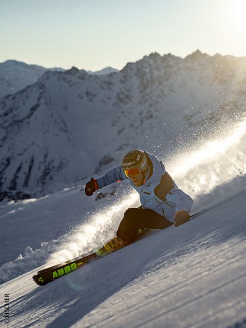 Un esquiador con chaqueta azul y casco desciende una ladera nevada al atardecer, levantando nieve en polvo, con picos de montañas escarpadas e iluminadas por el sol al fondo.