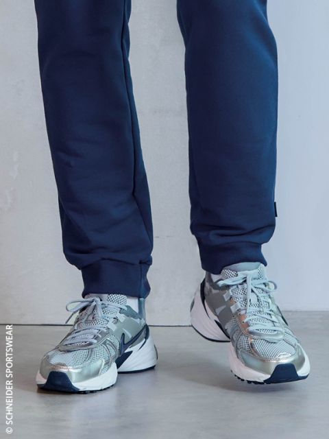 Una persona en pantalones de chándal azul marino y zapatillas de correr grises con suelas blancas está de pie en un interior sobre un suelo claro.