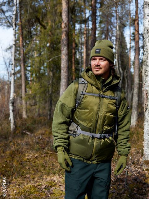 Un hombre con gorro, chaqueta y guantes de color verde oliva está de pie en un bosque iluminado por el sol y mira hacia un lado. Lleva una mochila y está rodeado de altos árboles y escasa maleza.
