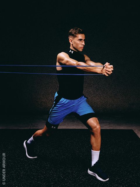 Un hombre en ropa deportiva realiza un ejercicio con banda de resistencia, de pie en posición de estocada y estirando las bandas con ambos brazos extendidos hacia adelante contra un fondo oscuro.