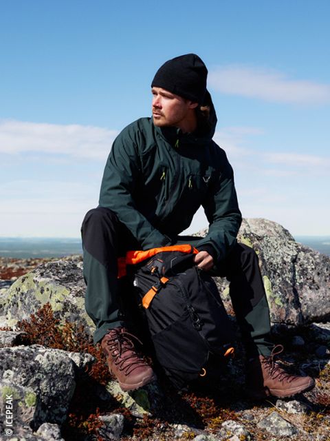 Un hombre con una chaqueta oscura, gorro negro y botas de montaña está sentado en un terreno rocoso al aire libre y lleva una mochila negra y naranja. El cielo es claro y azul al fondo.