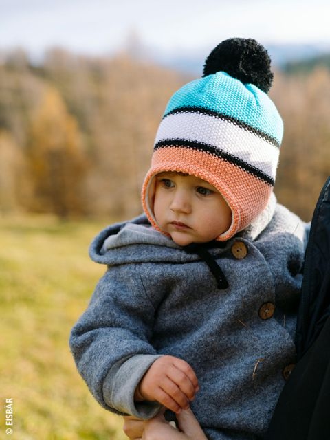 Un niño pequeño con un gorro de punto colorido y un abrigo gris es sostenido por un adulto al aire libre, con un fondo borroso de árboles de otoño y hierba.