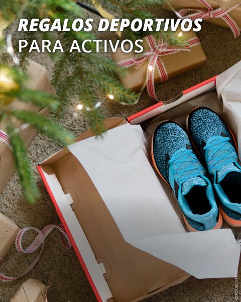 Caja de zapatos abierta con zapatillas deportivas azules, rodeada de regalos envueltos bajo un árbol de Navidad. El texto en la imagen dice: "Regalos deportivos para activos".