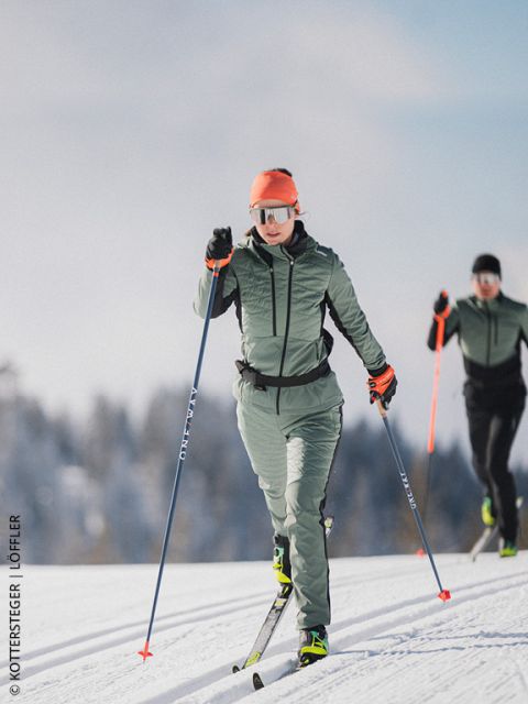 Dos esquiadores de fondo en una pista nevada, con equipo de deportes de invierno y gafas de sol, con árboles y cielo azul de fondo.