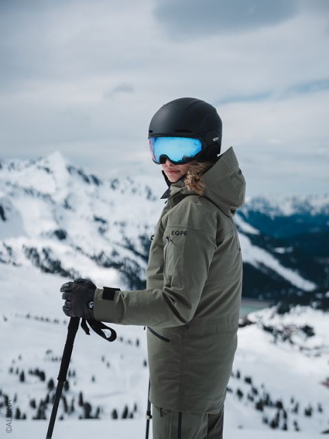 Un esquiador con chaqueta verde y casco negro está de pie en una ladera nevada, sosteniendo los bastones de esquí y mirando hacia atrás, con picos en el fondo.