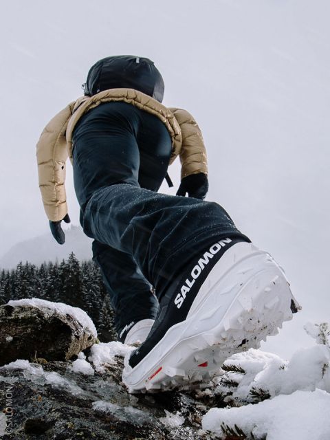 Una persona con ropa de invierno y zapatos Salomon camina cuesta arriba sobre la nieve, con el foco en la suela del zapato y un fondo de montaña nevada.