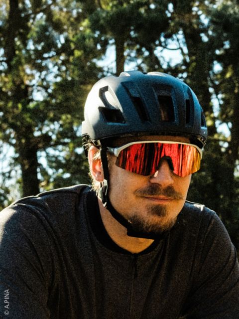 Un hombre con un casco de bicicleta negro, gafas de sol rojas reflectantes y una camisa oscura está de pie al aire libre con árboles al fondo.