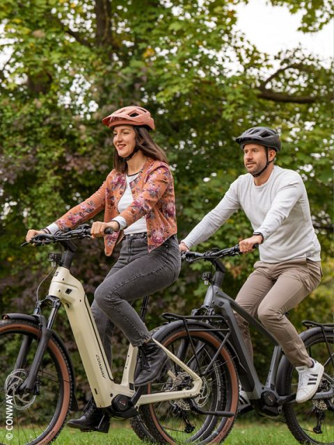 Una mujer y un hombre montan juntos en una bicicleta eléctrica en un parque verde, ambos llevan cascos y ropa informal, sonríen y disfrutan de la naturaleza.