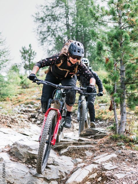 Dos ciclistas con cascos montan en sus bicicletas de montaña por un camino rocoso y boscoso, concentrándose y sorteando árboles y terrenos irregulares.
