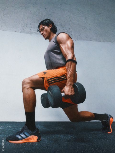 Un hombre musculoso en ropa deportiva realiza una zancada con mancuernas en un gimnasio y lleva zapatillas Adidas negras y naranjas.