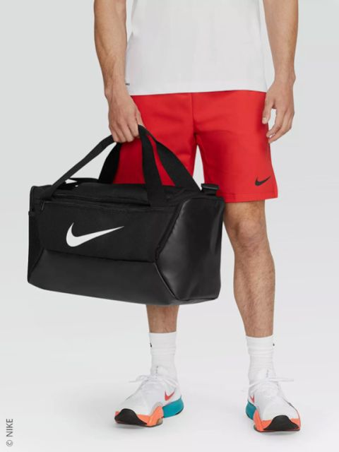 Una persona con pantalones cortos Nike rojos, camisa blanca y zapatillas blancas sostiene una bolsa de lona Nike negra con un logotipo Swoosh blanco.