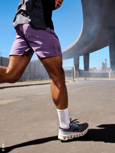 Persona caminando por una calle soleada bajo un paso elevado de una autopista, con pantalones cortos morados, calcetines blancos y zapatillas grises.