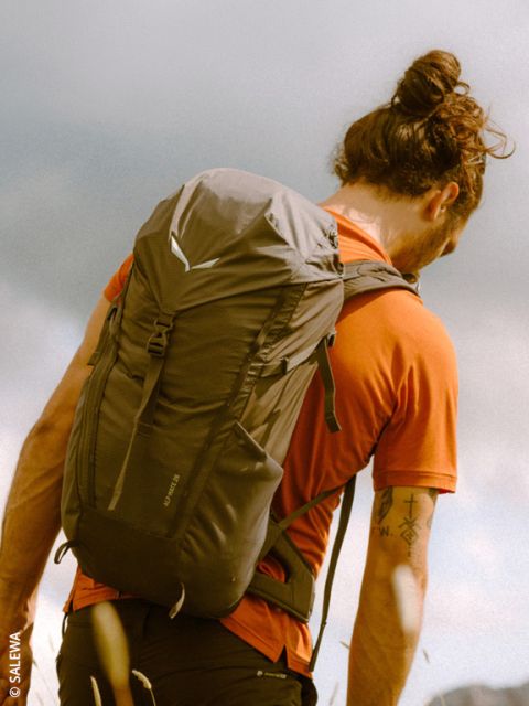 Una persona con un moño, una camisa naranja y una mochila está de pie al aire libre bajo un cielo nublado y mira a la distancia.