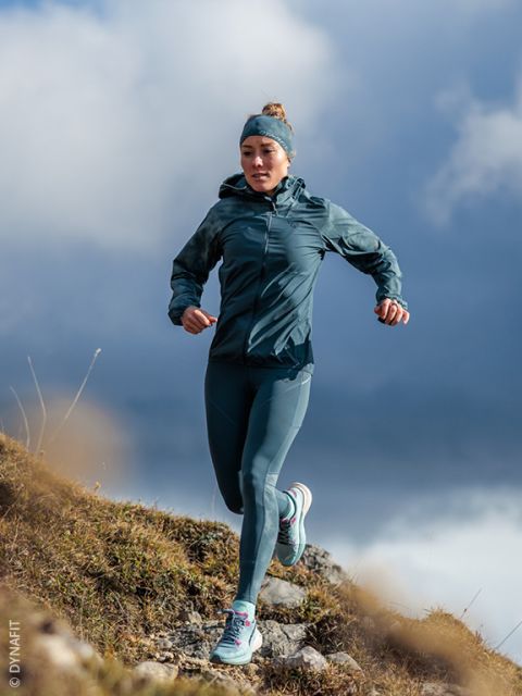 Una mujer con ropa deportiva turquesa corre cuesta abajo por un sendero rocoso, 
