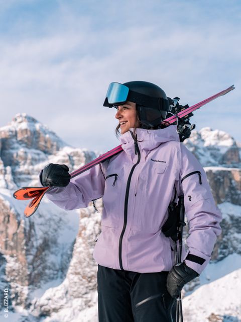 Una esquiadora con chaqueta lavanda y casco sonríe mientras lleva los esquís al 