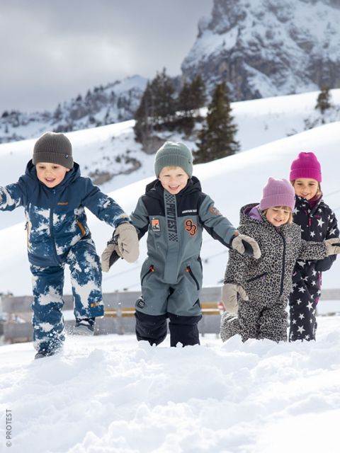 Cuatro niños con ropa de invierno juegan y sonríen en la nieve, con montañas y á