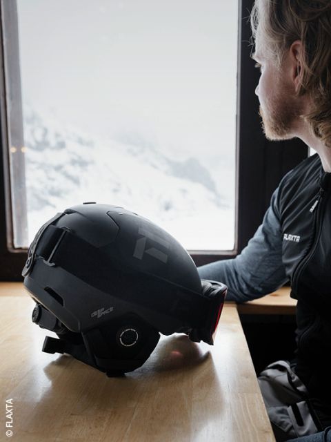 Una persona con cabello claro y barba está sentada en una mesa de madera junto a la ventana, mirando hacia las montañas nevadas. Un casco de esquí negro está delante de ellos sobre la mesa.